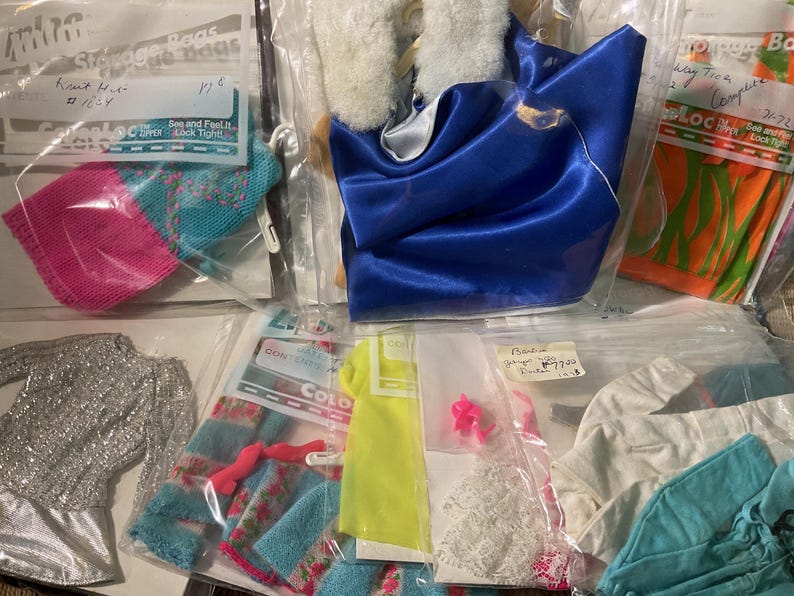 May include: A collection of doll clothing items packaged in individual clear plastic bags. The set includes a blue satin cape with a white faux fur collar, a silver glittery long-sleeve top, a pink and blue knit piece, and various other garments.