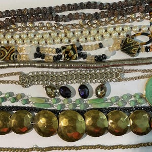 May include: A collection of necklaces featuring various materials and styles, including pearl, beaded, chain, and metal. The necklaces are arranged in rows, with the top row featuring a brown beaded necklace and the bottom row featuring a gold chain necklace with large, round, hammered metal beads.