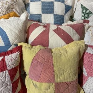 May include: A collection of square quilted pillows featuring various patchwork designs in colors including white, blue, red, yellow, and pink. The pillows display stitched patterns such as geometric blocks, star shapes, and fan motifs with textured fabric surfaces.