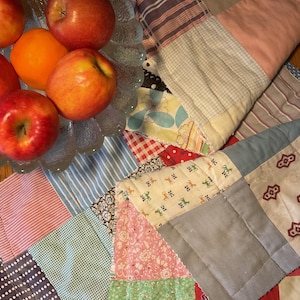 May include: A clear glass bowl filled with red apples and an orange sits atop a patchwork quilt. The quilt features various fabric patterns, including stripes, polka dots, and floral designs, in shades of blue, pink, and green.