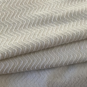 Puede incluir: Primer plano de una tela plegada de color blanquecino con un patrón de chevrón texturizado. La tela parece suave y tiene un brillo sutil. Los pliegues crean sombras, agregando profundidad a la imagen.