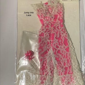 May include: A bright pink sleeveless dress with a white lace overlay, featuring a high neckline and a long skirt. The packaging includes the text Jump into Lace and Jump into Lace #1823. A small clear bag contains a pair of bright pink high-heeled shoes.