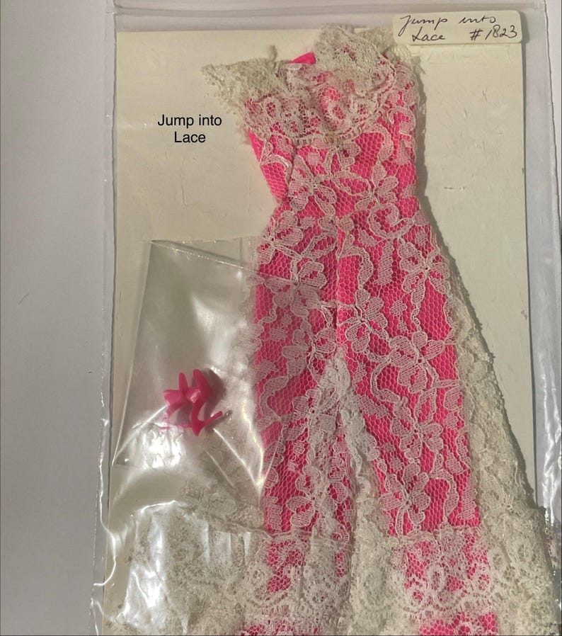 May include: A bright pink sleeveless dress with a white lace overlay, featuring a high neckline and a long skirt. The packaging includes the text Jump into Lace and Jump into Lace #1823. A small clear bag contains a pair of bright pink high-heeled shoes.