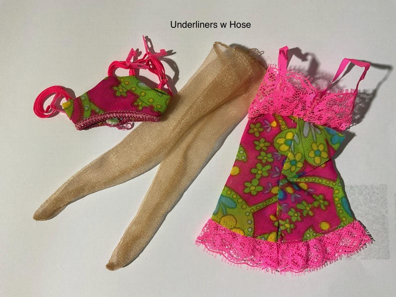 May include: A set of doll clothing labeled Underliners w Hose, featuring a pink and green floral patterned top with bright pink straps, a pair of sheer beige pantyhose, and a matching pink and green floral dress with bright pink lace trim.