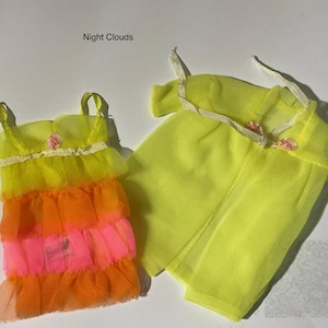 May include: A doll outfit labeled Night Clouds consisting of a sleeveless dress and a sheer robe. The dress features tiered ruffles in yellow, orange, and pink with a small pink flower accent. The yellow robe has thin straps and pink flower details.