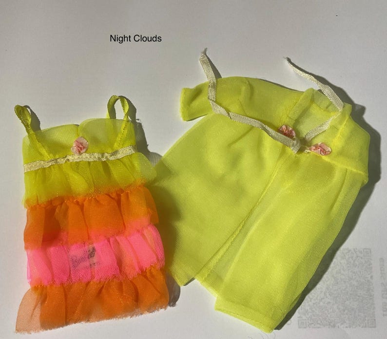 May include: A doll outfit labeled Night Clouds consisting of a sleeveless dress and a sheer robe. The dress features tiered ruffles in yellow, orange, and pink with a small pink flower accent. The yellow robe has thin straps and pink flower details.