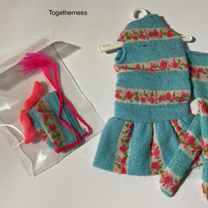 May include: A doll outfit set labeled Togetherness, featuring a light blue knit dress with pink floral stripes on a white background, matching knit leggings, a small knit accessory, and a pair of bright pink plastic doll shoes in a clear bag.