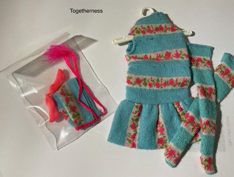 May include: A doll outfit set labeled Togetherness, featuring a light blue knit dress with pink floral stripes on a white background, matching knit leggings, a small knit accessory, and a pair of bright pink plastic doll shoes in a clear bag.