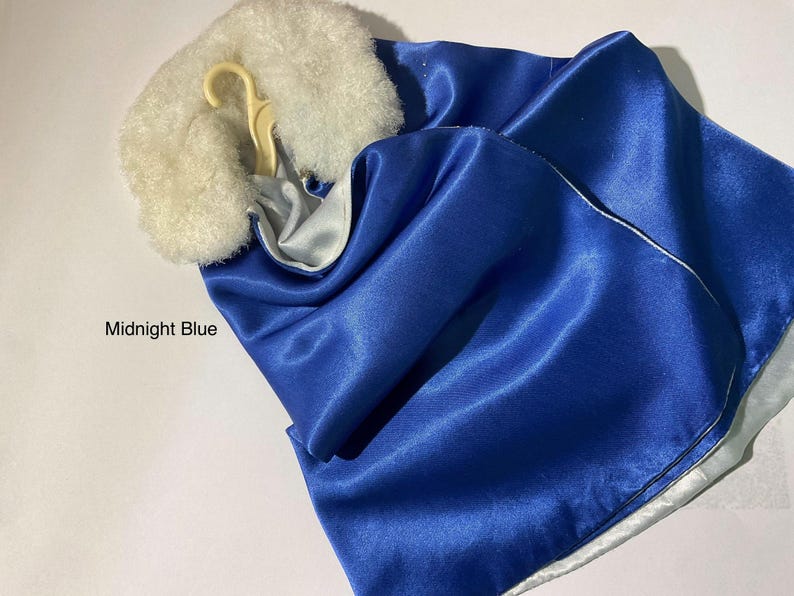 May include: A midnight blue satin cape featuring a white faux fur collar and a light-colored plastic hanger. The garment has a smooth, reflective surface and a white lining visible at the edges. The text Midnight Blue is printed on the left.