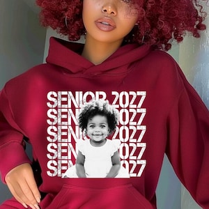 May include: A maroon hooded sweatshirt featuring a white graphic print on the chest. The design includes the repeated text SENIOR 2027 and a black and white photograph of a young child wearing a white dress in the centre of the text block.