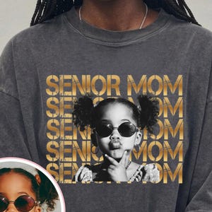 May include: A charcoal gray t-shirt featuring a repeating gold text graphic that reads SENIOR MOM. Centered is a black and white photograph of a child with pigtails wearing round sunglasses, with a circular inset showing the same child.