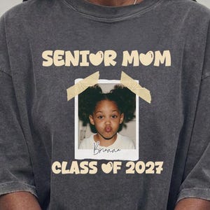 Puede incluir: Una camiseta de manga corta gris carb&oacute;n con el texto SENIOR MOM y CLASS OF 2027 en color crema. Centrada hay una foto de un ni&ntilde;o con coletas, enmarcada como una polaroid con cinta, con el nombre Brianna escrito en cursiva debajo del ni&ntilde;o.