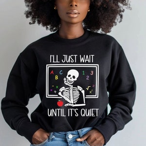 May include: Black sweatshirt with a white graphic of a skeleton teacher holding an apple and a chalkboard with the text "I'll just wait until it's quiet".