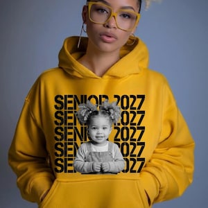 May include: A mustard yellow hooded sweatshirt featuring a black graphic print that repeats the text SENIOR 2027 multiple times. Centered on the chest is a black and white photograph of a young child with curly hair wearing overalls.