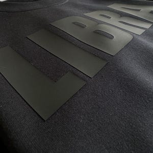 Op de afbeelding: Close-up van een donkergrijze sweatshirt met het woord "L BRA" in grote, zwarte blokletters over de borst. De letters zijn gemaakt van een plat, mat materiaal, wat een gedurfd contrast cre&euml;ert met de getextureerde stof van de sweatshirt.