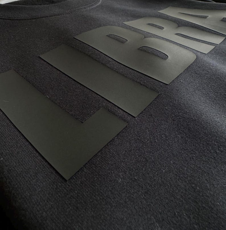 Op de afbeelding: Close-up van een donkergrijze sweatshirt met het woord "L BRA" in grote, zwarte blokletters over de borst. De letters zijn gemaakt van een plat, mat materiaal, wat een gedurfd contrast cre&euml;ert met de getextureerde stof van de sweatshirt.