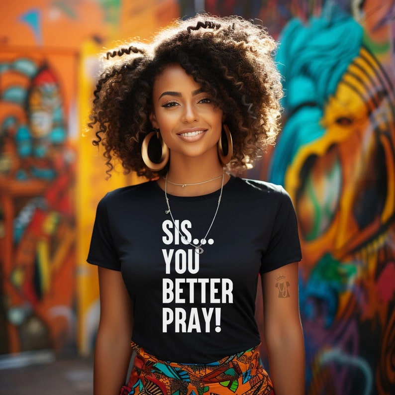 May include: A black t-shirt with the text "Sis... You Better Pray!" in white lettering. The model is wearing the shirt and has curly black hair and gold hoop earrings.
