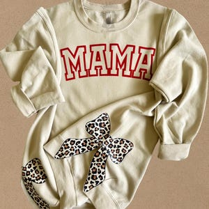 May include: A beige sweatshirt with the word "MAMA" printed in red on the front. The sweatshirt has a leopard print bow on the left sleeve.