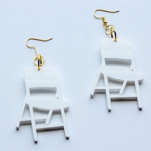 May include: A pair of white chair-shaped earrings with gold hooks. The chairs are made of a lightweight material and have a simple, modern design.