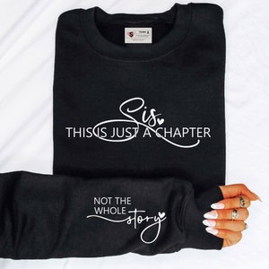 May include: Black sweatshirt with white text that reads "Sis, This Is Just A Chapter, Not The Whole Story." The sweatshirt has a crew neck and long sleeves. The sleeve is folded over, displaying the text on the cuff.