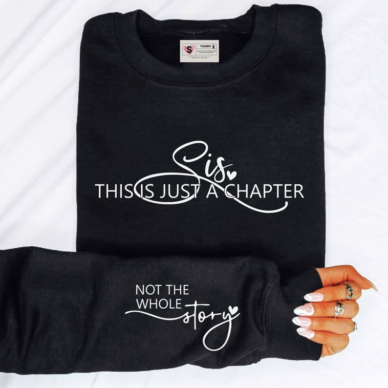 May include: Black sweatshirt with white text that reads "Sis, This Is Just A Chapter, Not The Whole Story." The sweatshirt has a crew neck and long sleeves. The sleeve is folded over, displaying the text on the cuff.