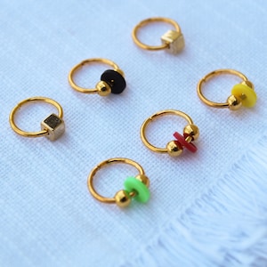 Puede incluir: Un conjunto de pendientes de aro dorados con varios abalorios de colores. Los abalorios incluyen un cubo dorado, un disco negro, un disco amarillo, una cruz roja y un disco verde. Se muestran sobre una superficie blanca.
