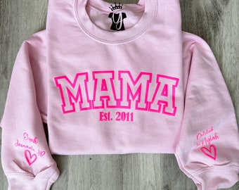 Op de afbeelding: Roze sweatshirt met het woord "MAMA" in roze omrande letters en de tekst "Est. 2011" eronder. De mouwen hebben roze harten en de namen "Elijah January" en "Delilah Nylah" in roze geschreven.