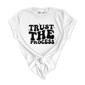 Puede incluir: Camiseta blanca con texto negro que dice "TRUST THE PROCESS" en una fuente retro.