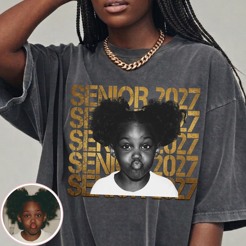 Op de afbeelding: Een antracietgrijs t-shirt met een herhalend goudkleurig tekstpatroon met de tekst SENIOR 2027. In het midden staat een zwart-wit fotoprint van een jong kind met haar in twee knotjes, gekleed in een wit t-shirt.