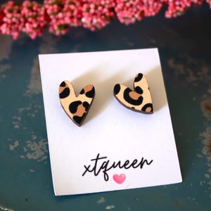 Könnte beinhalten: Ein Paar herzförmige Ohrringe aus Holz mit Leopardenmuster. Die Ohrringe befinden sich auf einer weißen Karte mit dem Text "xtqueen" und einem kleinen rosa Herz.