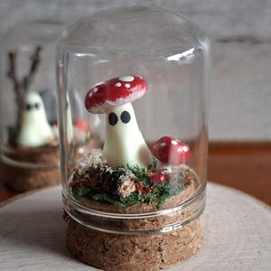 May include: A charming glass terrarium with a mushroom-shaped character, sporting a red and white cap and black eyes. The miniature landscape includes small red mushrooms and green moss, enclosed in a glass dome with a cork base. Another terrarium is visible in the background.