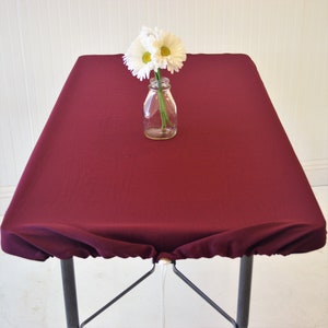 Peut inclure: Une nappe de table bordeaux avec un bord élastique ajusté recouvre une table pliante. Un vase en verre transparent avec des fleurs blanches est posé sur la table.