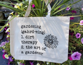 May include: A white canvas tote bag with black text that reads "gardening (gahrd-ning) 1. dirt therapy 2. the art of a gardener". A black and white line drawing of a daisy is to the right of the text.