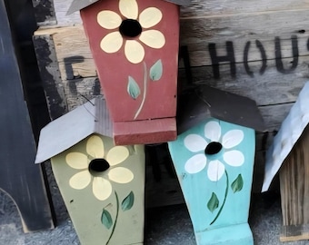 Peut inclure: Trois nichoirs en bois de couleurs rouge, vert et bleu, chacun orné d'une grande fleur peinte. Les nichoirs ont des toits gris foncé et sont placés sur un fond en bois rustique.