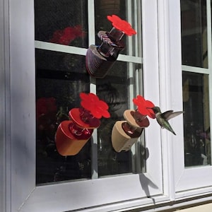 May include: Three hummingbird feeders attached to a window. Each feeder has a red flower-shaped top and a clear or tan base. A hummingbird is feeding from one of the feeders. The window frame is white.