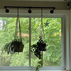 May include: Two hanging plants with green leaves in brown pots are suspended from a black metal pipe mounted above a window with a view of green trees.