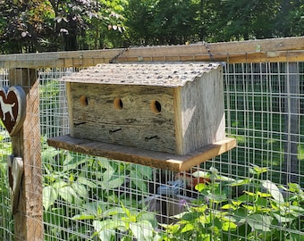 Könnte beinhalten: Ein verwittertes Vogelhaus aus Holz mit drei Nistfächern. Das Vogelhaus ist an einem Holzzaun mit einem Maschendrahtzaun im Hintergrund befestigt.