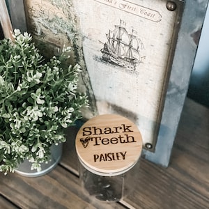 May include: A wooden jar with a lid that says "Shark Teeth Paisley" in a brown font. The jar is sitting on a wooden surface.
