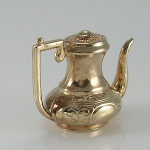 May include: A small, gold-colored teapot charm with a handle and spout. The teapot has a decorative lid and ornate detailing around the body. The charm is likely made of metal and is designed to be a pendant or charm.