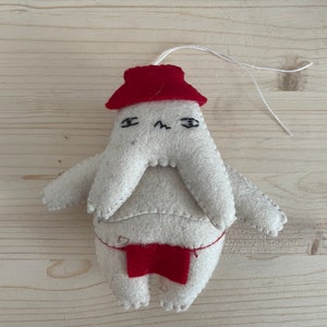 Op de afbeelding: Een wit vilten ornament met een rode muts en een rode broek. Het ornament heeft een eenvoudig gezicht met zwarte ogen en een frons.