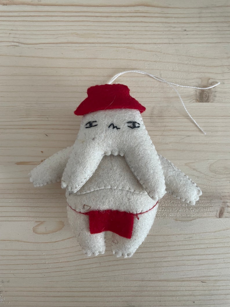 Op de afbeelding: Een wit vilten ornament met een rode muts en een rode broek. Het ornament heeft een eenvoudig gezicht met zwarte ogen en een frons.