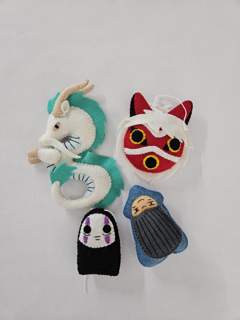 May include: Four felt ornaments featuring characters from a popular animated film. The ornaments include a white and teal dragon, a red and white mask, a black and white figure, and a blue figure with a gray blanket.