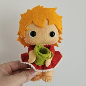 May include: A handmade felt doll with orange hair, large black eyes, and a light peach face. The doll wears a red dress with a white trim and holds a green bucket. The doll is a cute, whimsical design.