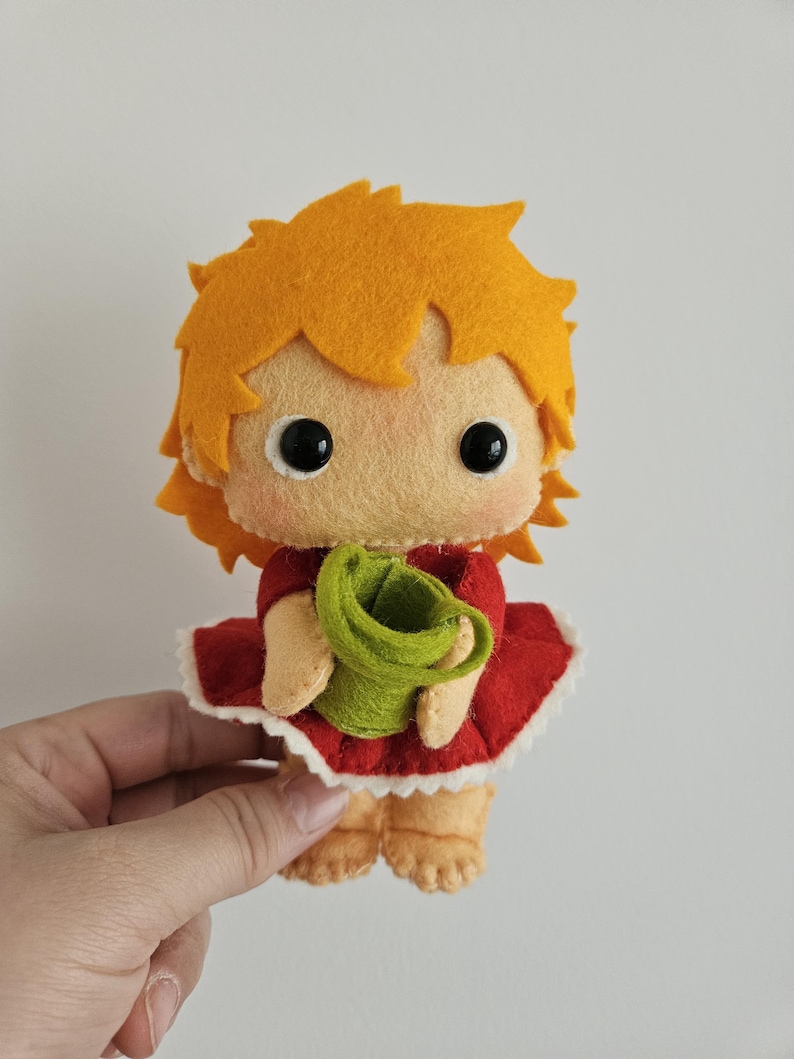 May include: A handmade felt doll with orange hair, large black eyes, and a light peach face. The doll wears a red dress with a white trim and holds a green bucket. The doll is a cute, whimsical design.