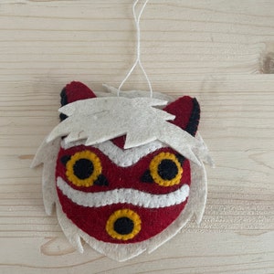 Op de afbeelding: Een vilten ornament van een rood en wit masker met zwarte ogen en een gele mond. Het masker heeft witte vacht rond de randen.