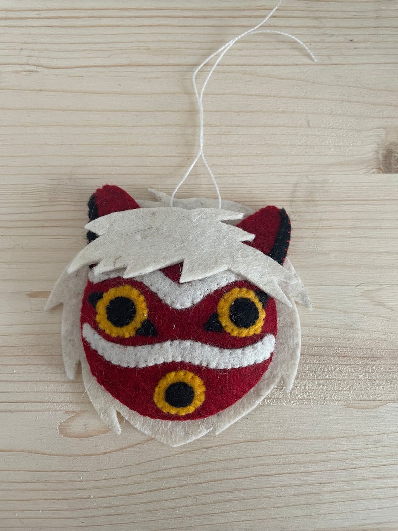 Op de afbeelding: Een vilten ornament van een rood en wit masker met zwarte ogen en een gele mond. Het masker heeft witte vacht rond de randen.