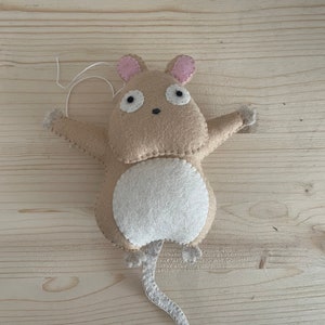 Op de afbeelding: Een beige viltmuis met roze oren, een witte buik en een lange staart. De muis hangt aan een touwtje.