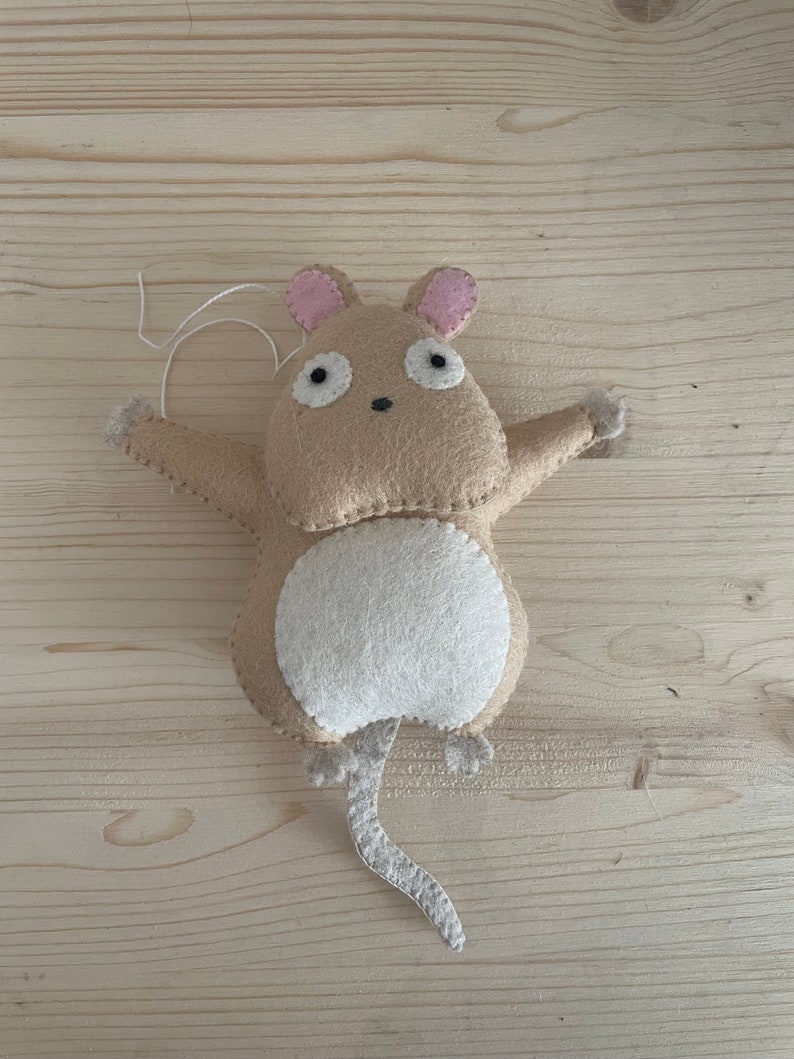 Op de afbeelding: Een beige viltmuis met roze oren, een witte buik en een lange staart. De muis hangt aan een touwtje.
