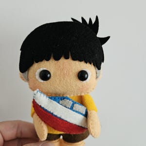 May include: A handmade felt doll of a boy with black hair, wearing a yellow shirt and brown shorts. The doll is holding a small boat with red, white, and blue details. The doll has black button eyes and is made of soft felt.