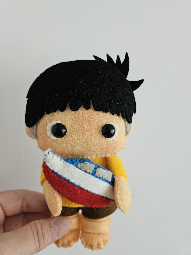 May include: A handmade felt doll of a boy with black hair, wearing a yellow shirt and brown shorts. The doll is holding a small boat with red, white, and blue details. The doll has black button eyes and is made of soft felt.